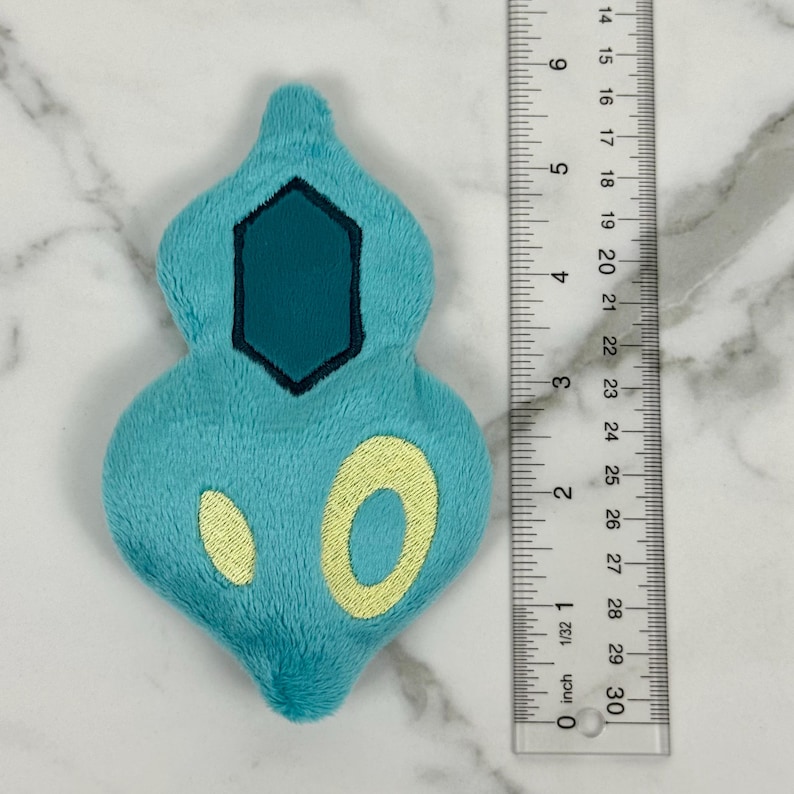 May include: A plush, turquoise-colored figure with a dark teal hexagon and yellow circular accents. The figure is approximately 3 inches long, as measured by the ruler in the image. The plush has a soft, textured appearance.