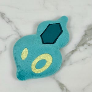 May include: A plush toy in a unique shape, primarily turquoise with yellow and dark teal accents. The toy features a central dark teal hexagon and two yellow oval shapes. The plush material gives it a soft appearance.