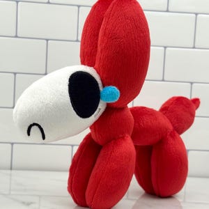 Puede incluir: Un juguete de peluche rojo con forma de perro, al estilo de un animal de globo. Tiene una cabeza blanca con un ojo negro y un pequeño detalle azul. El juguete está hecho de tela suave.