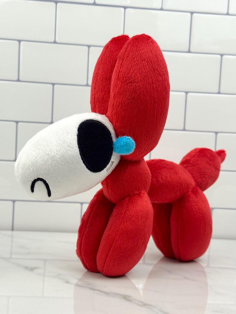 May include: A plush toy dog in a balloon animal style. The dog is primarily red with a white head, black eye, and a small blue accent. The toy is made of soft fabric and has a curved tail.