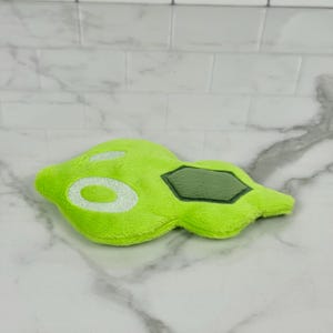 May include: A lime green plush toy in an abstract shape, featuring white embroidered details and a dark green geometric patch. The toy is made of soft, fuzzy material and rests on a marble surface.