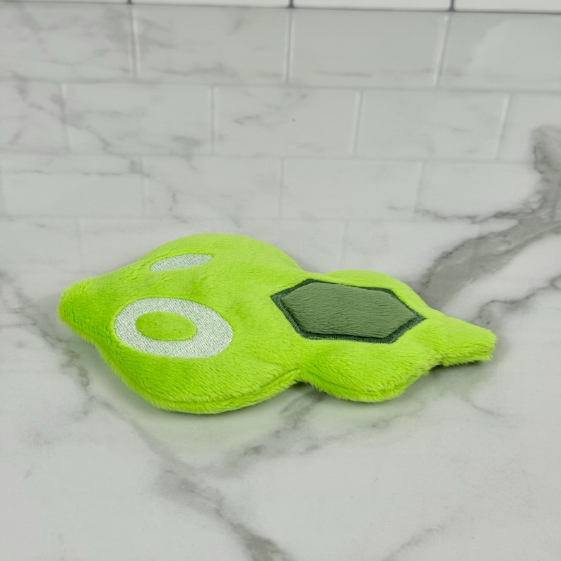 May include: A lime green plush toy in an abstract shape, featuring white embroidered details and a dark green geometric patch. The toy is made of soft, fuzzy material and rests on a marble surface.