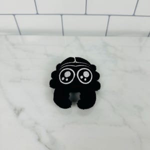 May include: A black plush spider with white embroidered eyes. The spider has eight legs and a round body.