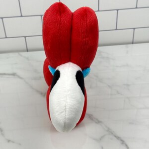 May include: A plush toy featuring a red head with two large, heart-shaped sections. The toy has a white face with black accents and a blue collar. The toy is designed to resemble a character.