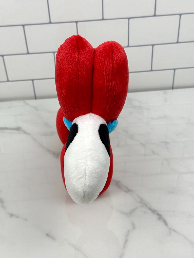 May include: A plush toy featuring a red head with two large, heart-shaped sections. The toy has a white face with black accents and a blue collar. The toy is designed to resemble a character.