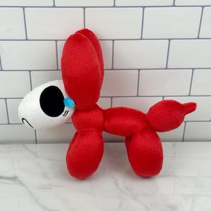 May include: A plush toy in the shape of a red balloon dog with a white head and black eyes. The toy has a small blue accent near the nose and is made of soft, fuzzy material. The dog is standing on a white and gray surface.