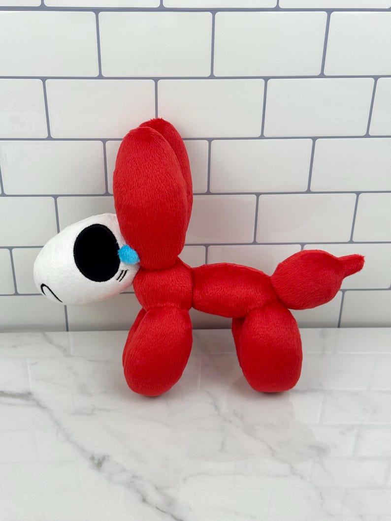 May include: A plush toy in the shape of a red balloon dog with a white head and black eyes. The toy has a small blue accent near the nose and is made of soft, fuzzy material. The dog is standing on a white and gray surface.