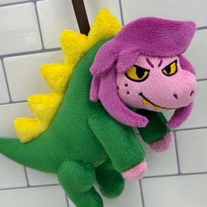 May include: A plush dinosaur toy with a green body, yellow spines, and a pink face and hair. The dinosaur has yellow eyes and a brown strap attached to the top. The toy is made of soft, plush material.