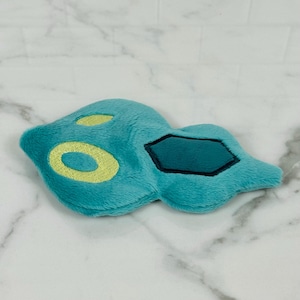 May include: A plush toy in the shape of a teal-colored creature with yellow and black accents. The toy has a soft, fuzzy texture and a unique, stylized design. The toy is lying on a white marble surface.