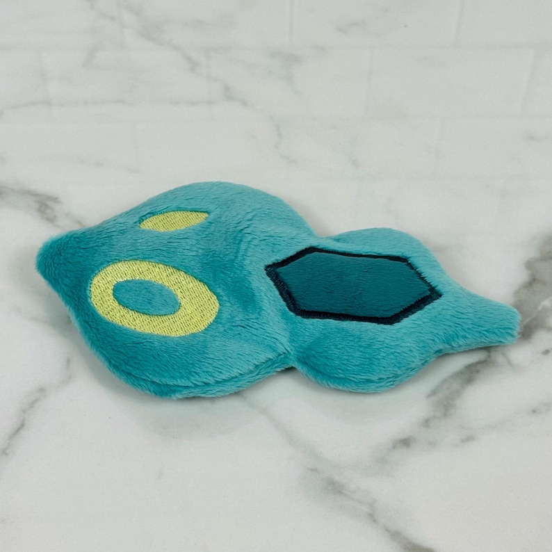 May include: A plush toy in the shape of a teal-colored creature with yellow and black accents. The toy has a soft, fuzzy texture and a unique, stylized design. The toy is lying on a white marble surface.