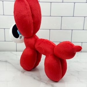 May include: A plush toy in the shape of a red balloon dog. The toy has large, floppy ears and a white and black eye with a blue iris. The dog is made of soft, plush material.