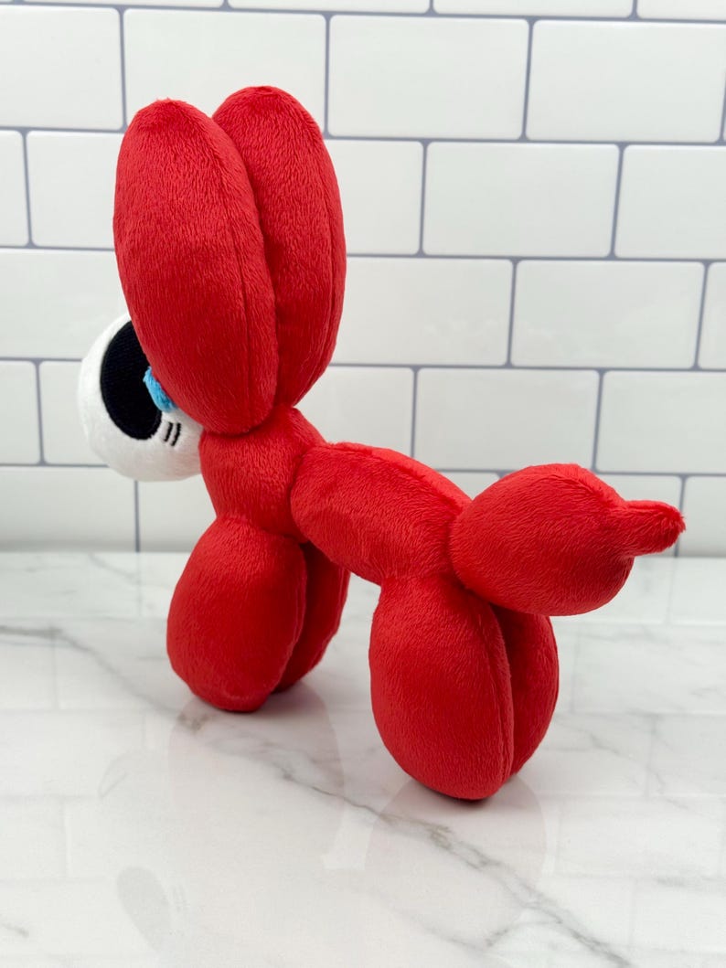 May include: A plush toy in the shape of a red balloon dog. The toy has large, floppy ears and a white and black eye with a blue iris. The dog is made of soft, plush material.