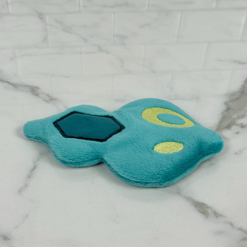 May include: A plush toy in the shape of a stylized blue creature with yellow and dark blue accents. The toy has a soft, fuzzy texture and is lying on a white marble surface.