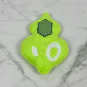 May include: A lime green plush toy in a unique shape, featuring a dark green hexagon and white embroidered details. The plush has a soft, fuzzy texture and is set against a marble background.