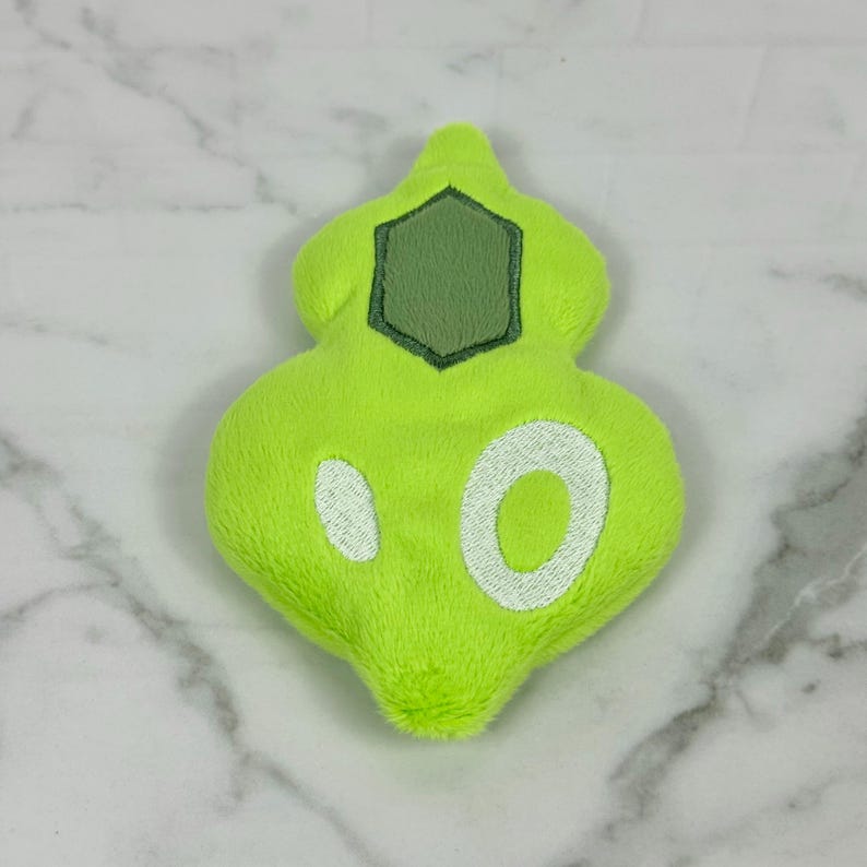 May include: A lime green plush toy in a unique shape, featuring a dark green hexagon and white embroidered details. The plush has a soft, fuzzy texture and is set against a marble background.
