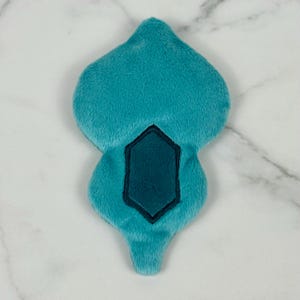 May include: A plush, teal-colored, teardrop-shaped item with a darker teal, hexagonal design in the center. The item is made of soft, fuzzy material and is likely a decorative or novelty item.