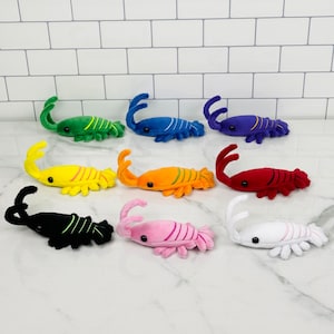 Könnte beinhalten: Ein Set aus zehn Plüschtieren in Form von Hummern in verschiedenen Farben, darunter Rot, Blau, Grün, Gelb, Schwarz, Weiß und Pink. Die Hummer liegen alle in einer Reihe auf einer weißen Oberfläche.