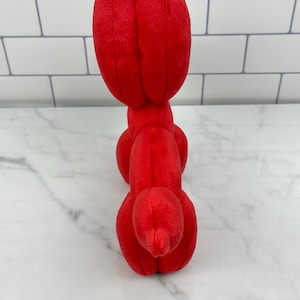 May include: A bright red plush toy shaped like a balloon animal. The toy is made of soft, fuzzy material and features a rounded head and body with elongated sections. The toy is positioned upright against a white and gray background.