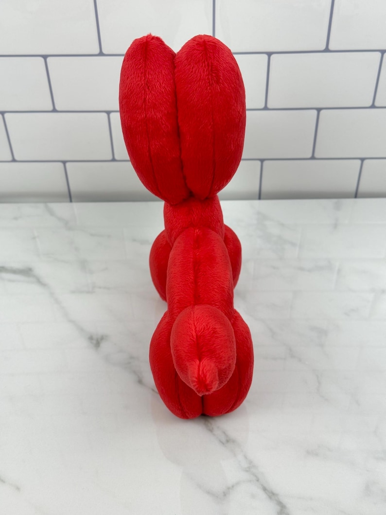 May include: A bright red plush toy shaped like a balloon animal. The toy is made of soft, fuzzy material and features a rounded head and body with elongated sections. The toy is positioned upright against a white and gray background.