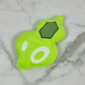 May include: A plush, lime green toy with a unique, organic shape. It features a dark green, hexagonal patch and two white, oval-shaped accents. The toy is made of a soft, fuzzy material, and is likely a children's toy or a decorative item.