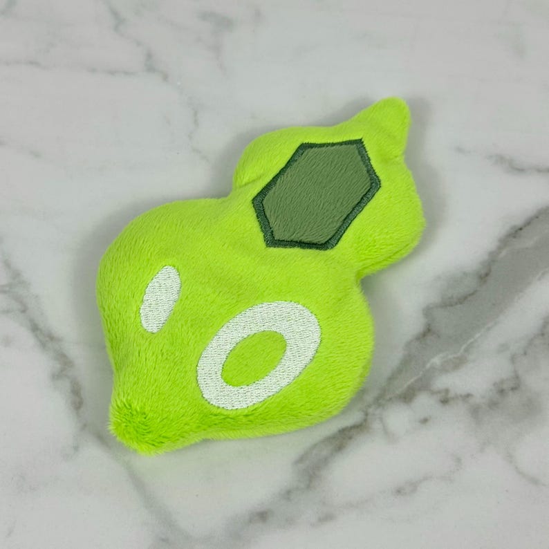 May include: A plush, lime green toy with a unique, organic shape. It features a dark green, hexagonal patch and two white, oval-shaped accents. The toy is made of a soft, fuzzy material, and is likely a children's toy or a decorative item.