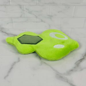 May include: A lime green plush toy with a unique shape, featuring a darker green, hexagonal patch and white circular accents. The soft, textured surface suggests a cuddly item, possibly a pet toy or decorative piece.
