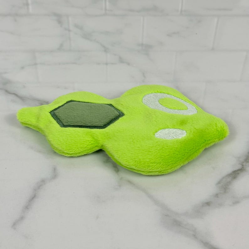 May include: A lime green plush toy with a unique shape, featuring a darker green, hexagonal patch and white circular accents. The soft, textured surface suggests a cuddly item, possibly a pet toy or decorative piece.