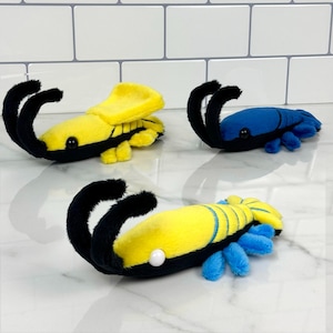 May include: Three plush toys shaped like shrimp. One is yellow with black accents, one is blue with black accents, and one is yellow with black and blue accents.