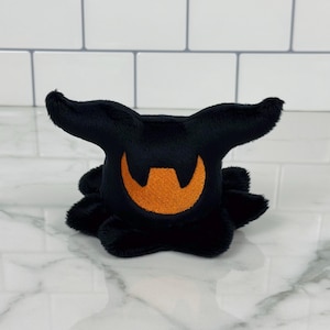 May include: A black plush toy with a smiling orange face and two pointed ears. The toy has a round body with several small, rounded appendages. The plush is sitting on a white surface.