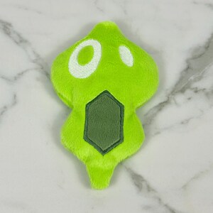 May include: A lime green plush toy with a unique shape. It features two white circular accents and a dark green, geometric design at the bottom. The toy is made of a soft, fuzzy material.