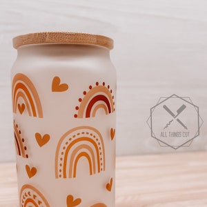 May include: A white ceramic jar with a wooden lid, featuring a repeating pattern of orange, red, and yellow rainbows and hearts. The jar is decorated with a boho-style design.