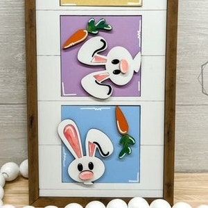 May include: A wooden framed wall decor with three bunny rabbits and carrots. Each bunny is on a different colored square. The squares are yellow, purple, and blue. The frame is brown.