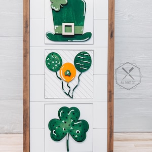 May include: A framed wood sign with three painted wooden decorations. The top decoration is a green leprechaun hat. The middle decoration is three green balloons with a yellow balloon in the center. The bottom decoration is a green shamrock. The sign is on a small wooden easel.