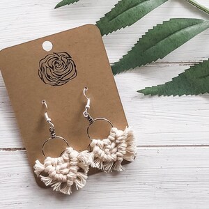 May include: A pair of white macrame earrings with silver hoops. The earrings are hanging from a brown card with a rose design.