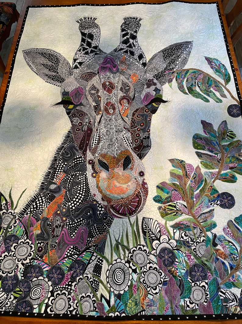 May include: A close-up of a colorful, patterned quilt featuring a giraffe's head and neck. The giraffe is surrounded by a variety of flowers and leaves, all in a similar style. The quilt is made of fabric scraps and is a unique and creative piece of art.