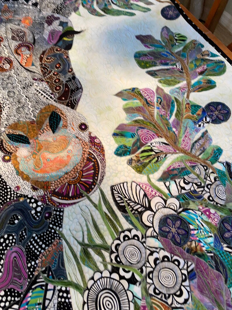 May include: A colorful quilt with a white background featuring a large, abstract floral design. The quilt is made of various fabrics and textures, including black and white polka dots, green leaves, and purple flowers.