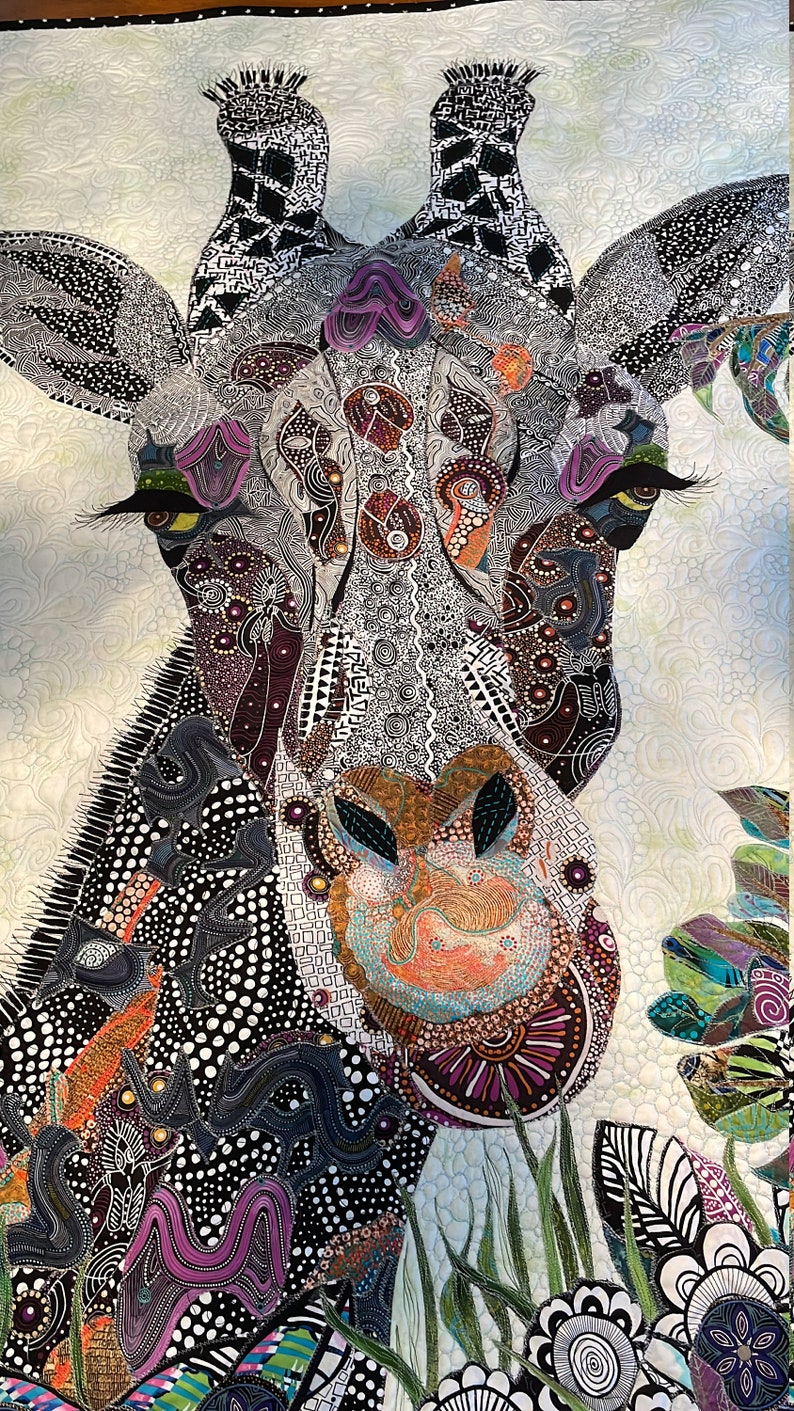 May include: A close-up of a colorful, patterned quilt featuring a giraffe's head. The giraffe's face is detailed with black and white lines, and its eyes are outlined in black with yellow pupils. The quilt is made of various fabrics, including black, white, purple, orange, and green.