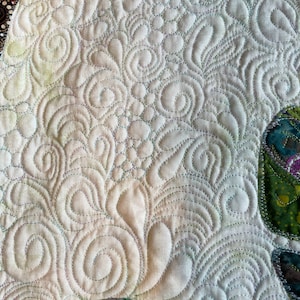May include: Close-up of a white fabric with a detailed, intricate, swirling, quilted pattern. The quilting is done in a light green thread. The fabric is textured and has a slight sheen.