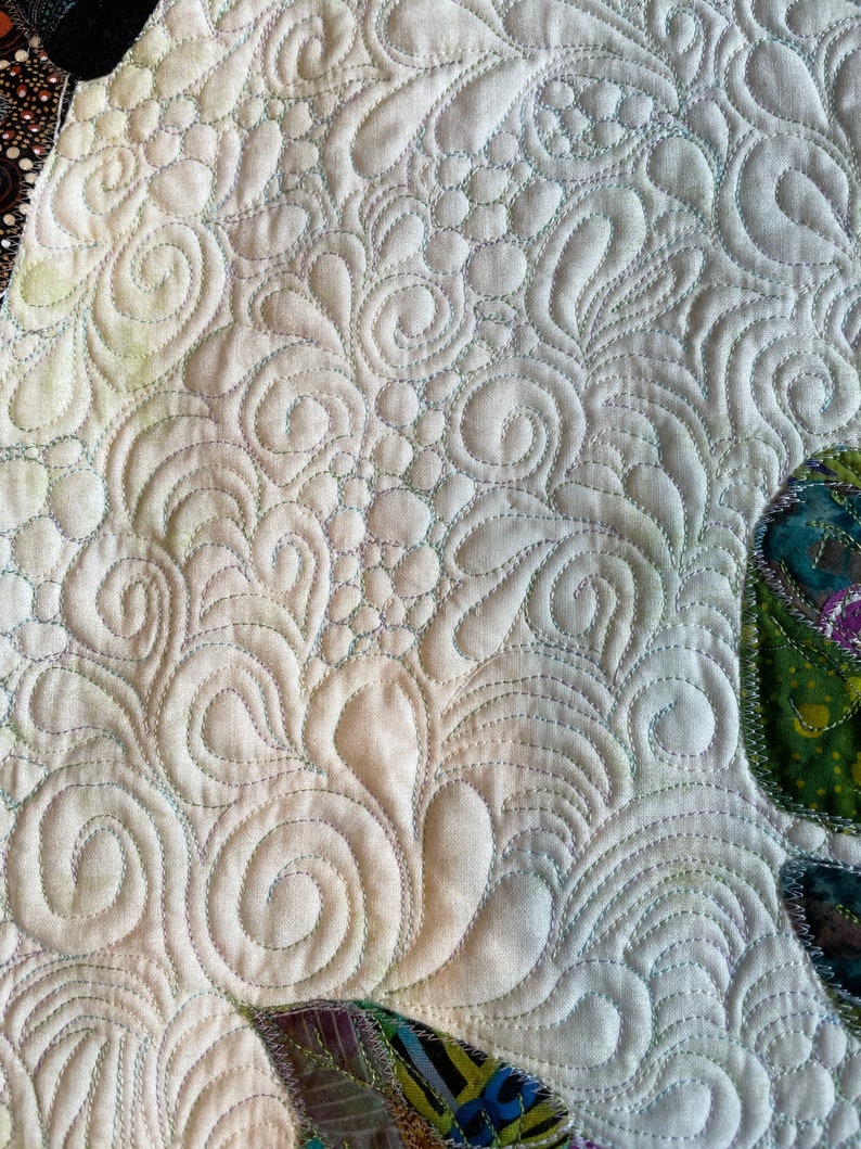 May include: Close-up of a white fabric with a detailed, intricate, swirling, quilted pattern. The quilting is done in a light green thread. The fabric is textured and has a slight sheen.