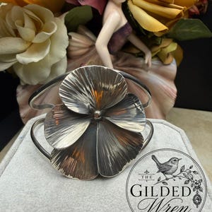 May include: A silver cuff bracelet featuring a large, dimensional flower centerpiece with textured, radiating petals. The metal has a darkened, oxidized finish in the recessed areas, highlighting the detailed, fan-like structure of the floral design.