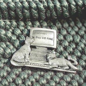 May include: A silver-toned metal pin featuring two cats interacting with a computer. One cat sits upright beside the monitor, while the other lies across the keyboard. The screen displays the text: Memo: * Buy cat food in raised lettering.