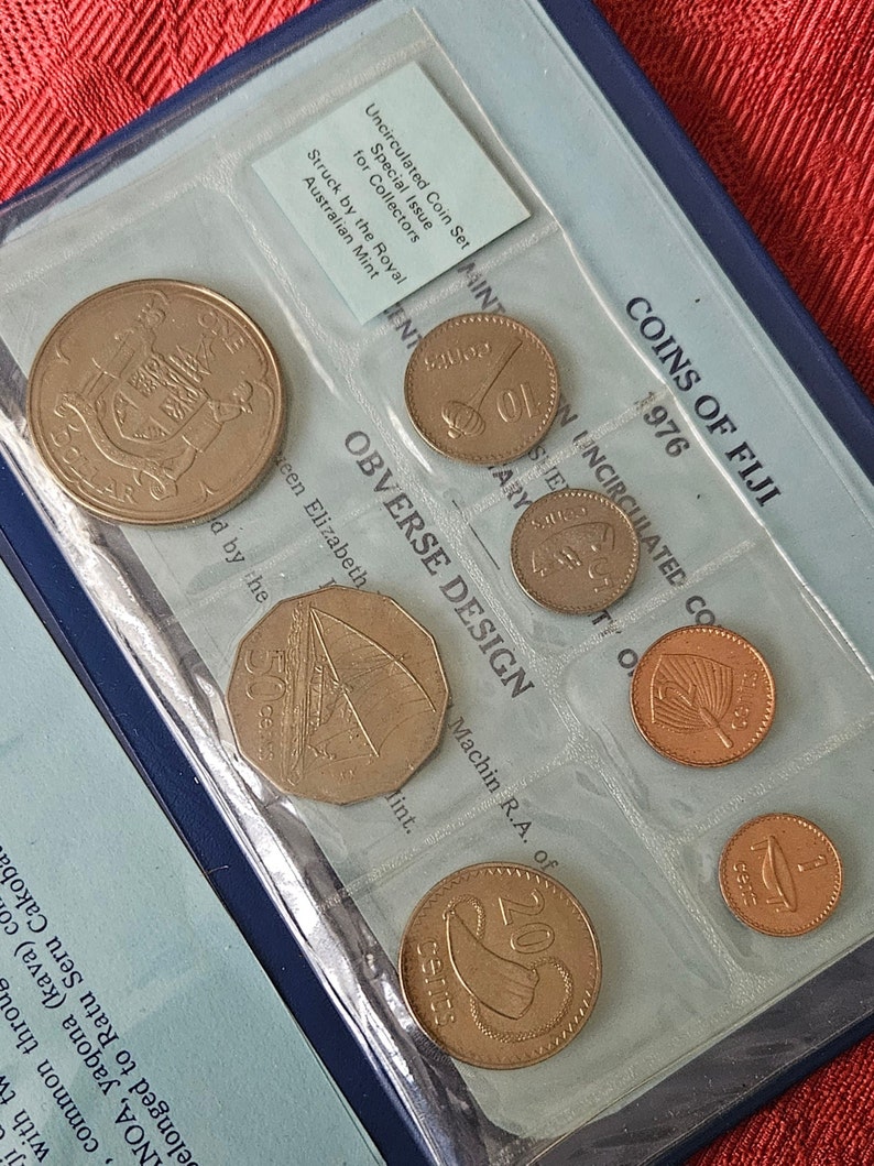 May include: A blue coin collection folder with a clear plastic insert showing a set of uncirculated Fijian coins. The coins are in denominations of 1, 5, 10, 20, and 50 cents. The folder has the text "COINS OF FIJI 1976" printed on the front.