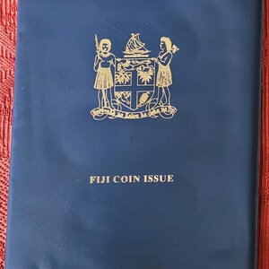 May include: A blue leather coin album with gold lettering that reads "FIJI COIN ISSUE" and a gold crest with two figures and a ship.