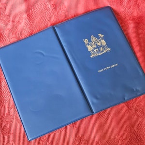 May include: A blue coin album with a gold emblem of the Fijian coat of arms and the text "FIJI COIN ISSUE".