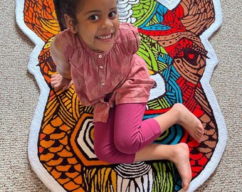 May include: A child sits on a colorful, patterned rug shaped like a camel. The rug has a white background with brown, orange, yellow, green, blue, and red patterns. The child is wearing a pink shirt and pants.