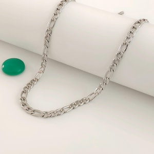 Può includere: Una collana a catena Figaro color argento. La collana presenta un motivo di maglie allungate e rotonde alternate. La catena è esposta su una superficie bianca con una pietra verde a forma ovale.
