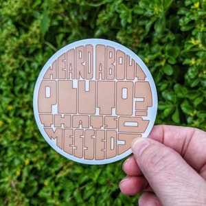 May include: A white round sticker with brown text that reads "Heard About Pluto? That's So Messed Up".