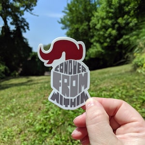 Pode incluir: Um autocolante com um design de capacete de cavaleiro estilizado com as palavras "CONQUER FROM WITHIN" em letras cinzentas. O capacete tem uma pluma vermelha. O autocolante é segurado contra um fundo de relva verde e árvores.