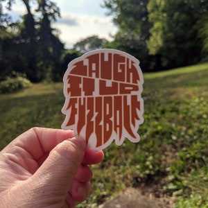 May include: A white sticker with an orange design that says "Laugh it up Fuzzball".