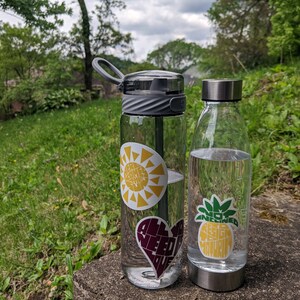 Puede incluir: Dos botellas de agua con pegatinas coloridas. La botella de la izquierda tiene una pegatina de sol amarilla con el texto "Feel Some Sun", una pegatina de coraz&oacute;n morado con el texto "All You Need Is Love", y una pegatina en blanco y negro con un dise&ntilde;o de sol y luna. La botella de la derecha tiene una pegatina de pi&ntilde;a verde y amarilla con el texto "Not Inclined To Design Maturity".